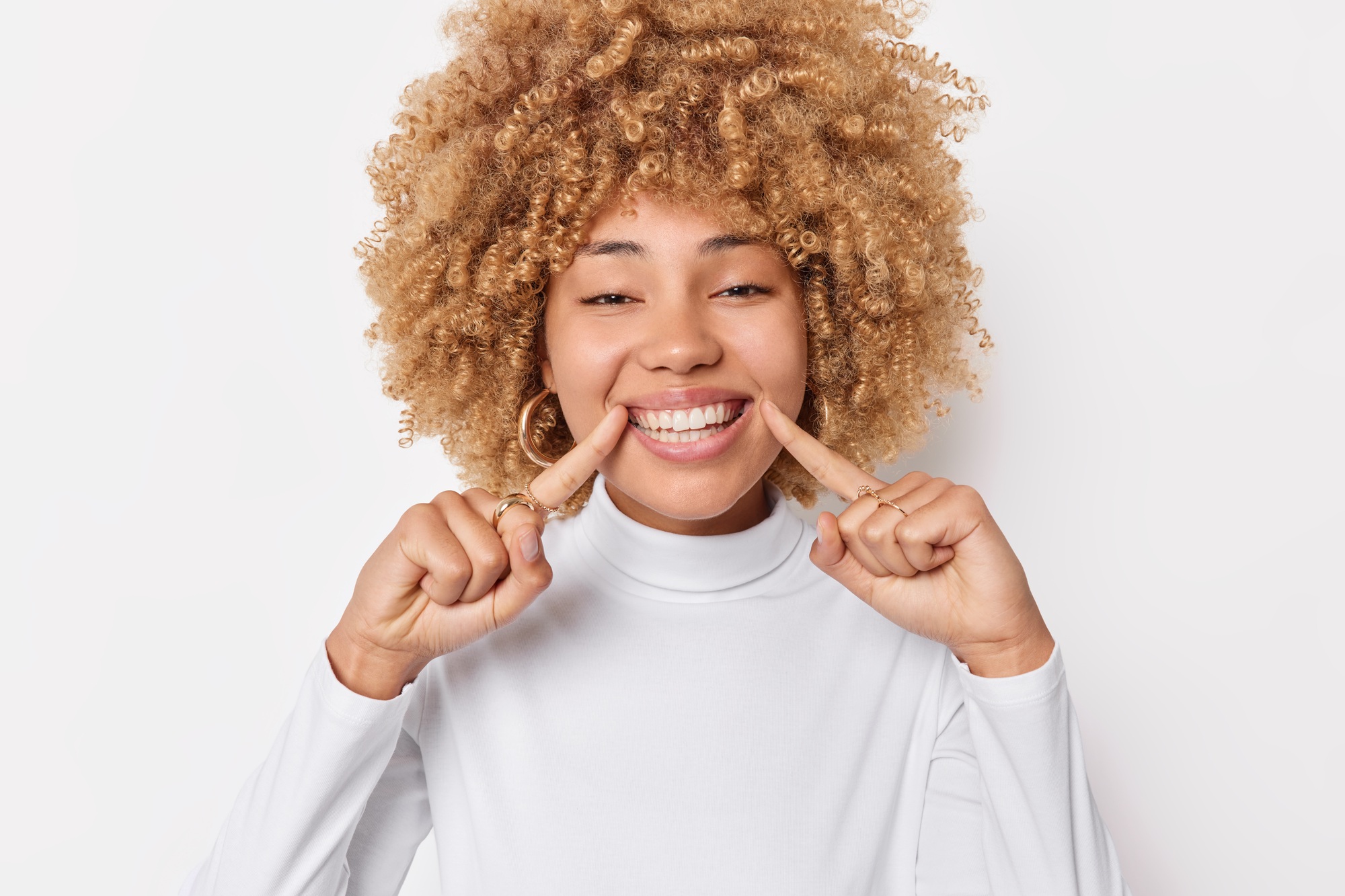 Look at my perfect orthodontic smile. Positive woman with curly bushy hair cheerful expression point