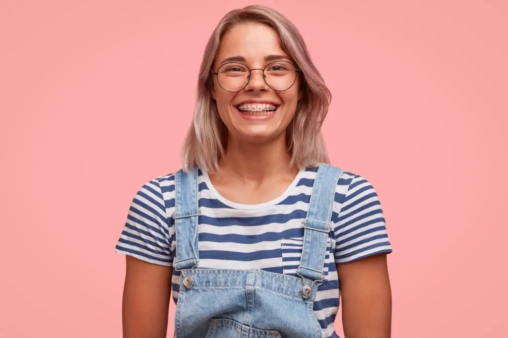 Positive glad smiling female teenager with braces on teeth, wears stylish denim overalls and striped